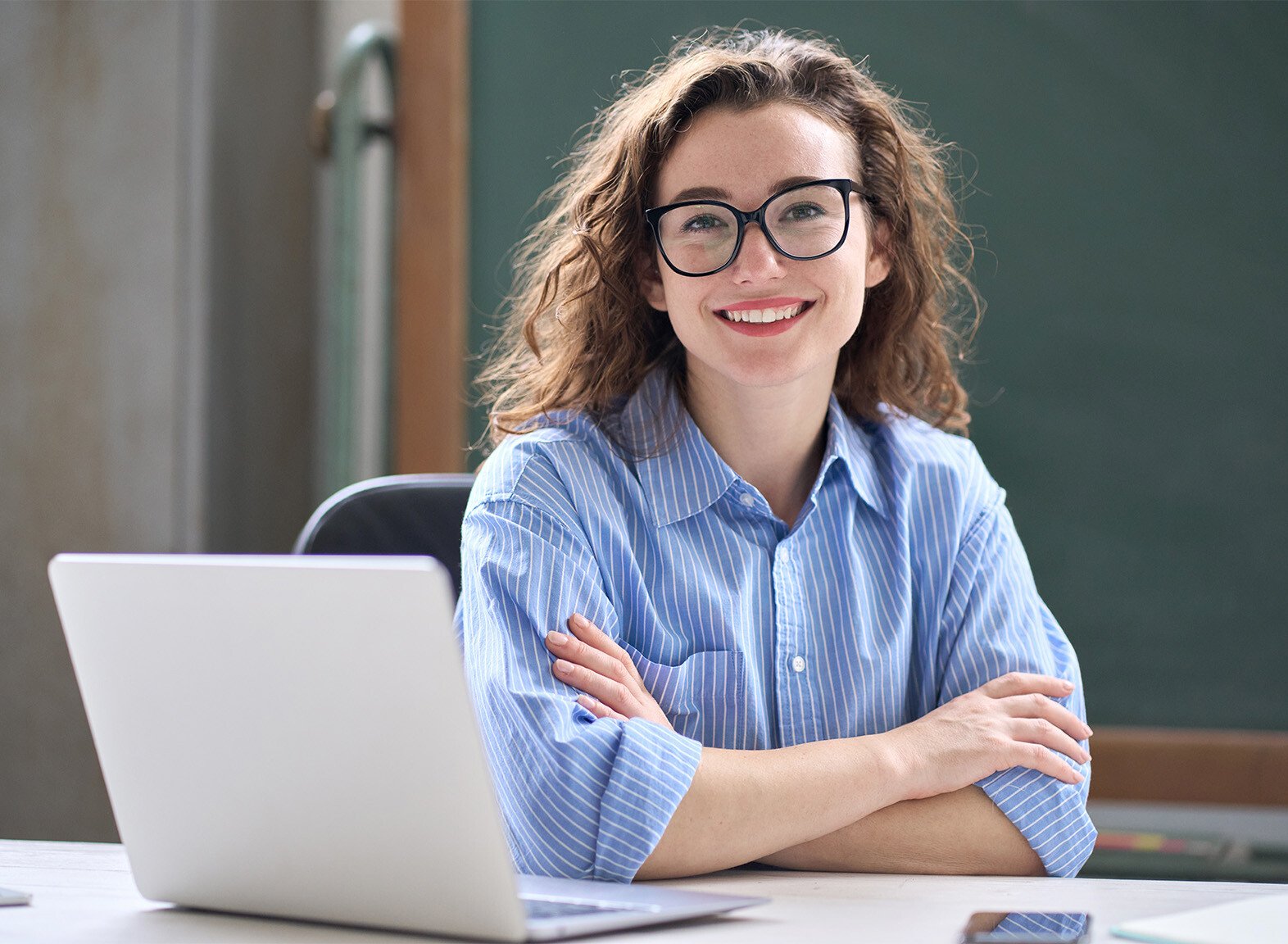 Happy female teacher sat infront of a laptop, looking into the camera with her arms crossed