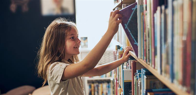 Happy primary school aged girl reaching for a book in a library