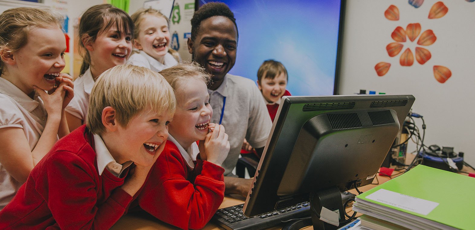 Primary aged school students gathered around the teachers computer, smiling